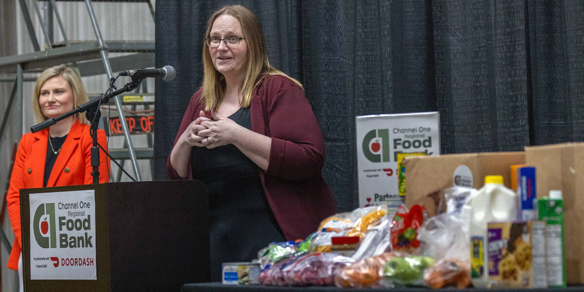Two Women Stand Near A Podium At A Food Bank Event. Various Food Items Are Displayed On A Table In The Foreground.