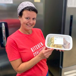 Channel One Regional Food Bank Kitchen Coalition Person In A Red Kitchen Coalition Shirt Holds A Container Of Packaged Food In A Kitchen Setting.