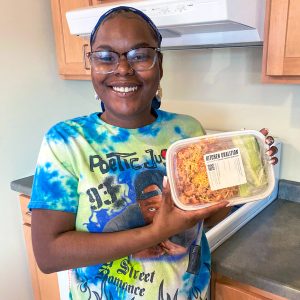 Channel One Regional Food Bank Kitchen Coalition3 Person Smiling In Kitchen, Holding A Meal Container Labeled Kitchen Coalition.