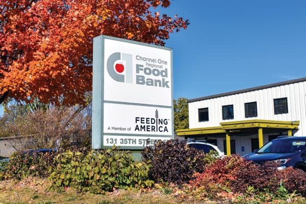 Channel One Regional Food Bank And Food Shelf 2024 Sign For Channel One Regional Food Bank, A Feeding America Member, In Front Of A Building, Surrounded By Autumn Trees And Parked Cars.
