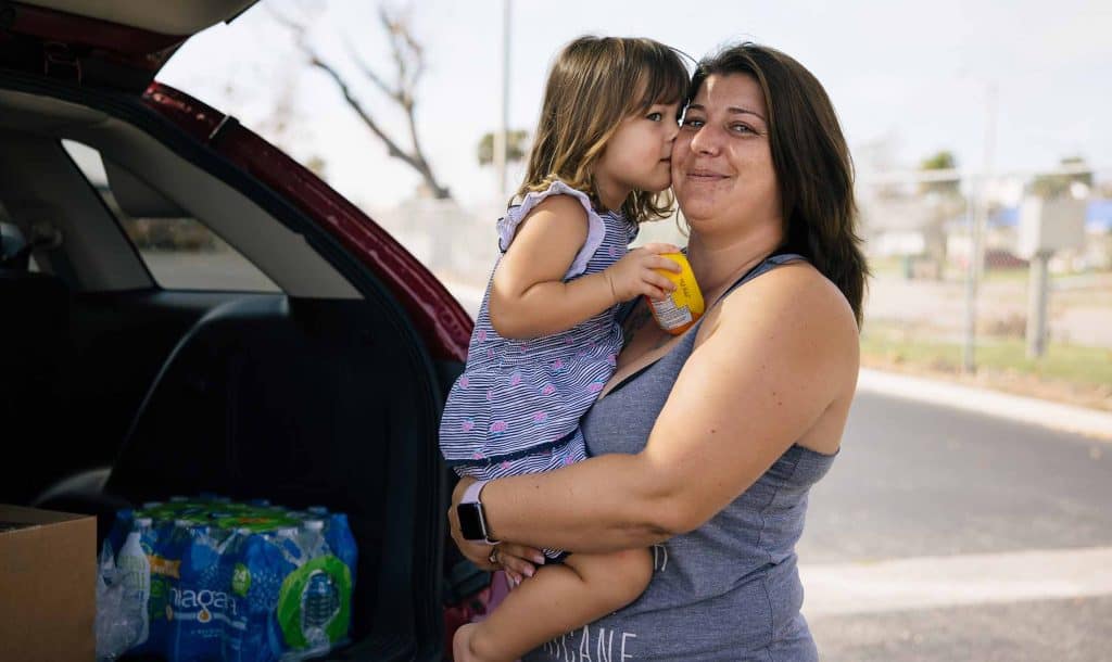 Mother And Young Daughter Standing Near Open Trunk Of Car With Food Inside