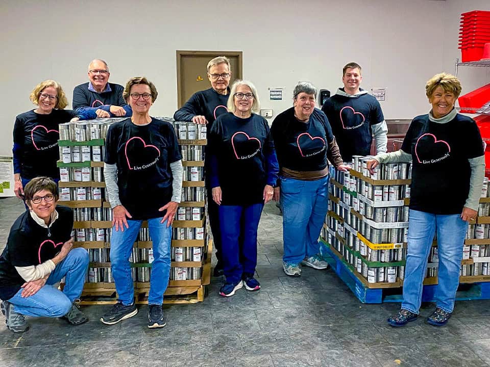 Channelonegetinvoledcorporationsandbusinesses2025 A Group Of People Wearing Matching T-Shirts Stands Around Pallets Stacked With Canned Goods In A Room.