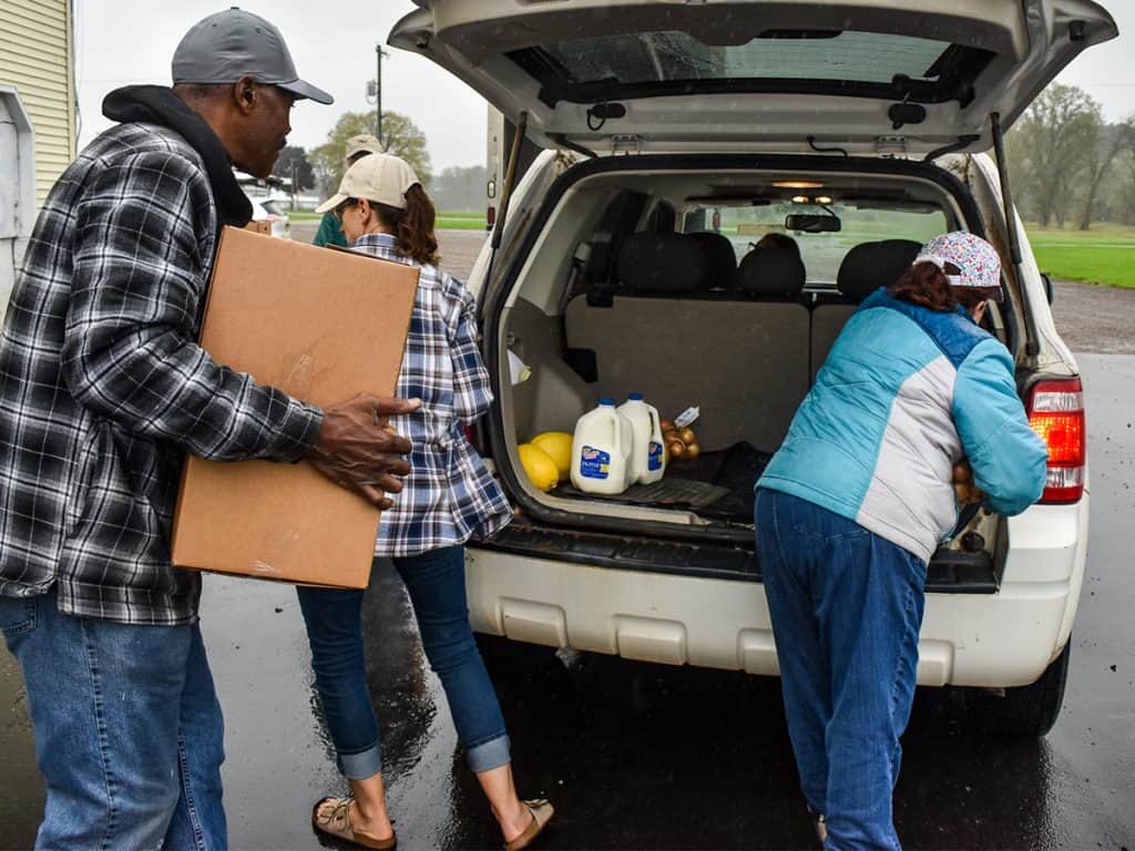 Channelonemobilepantries2025 Three People Load A White Vehicle With A Cardboard Box, Jug Of Milk, And Other Groceries On A Rainy Day.