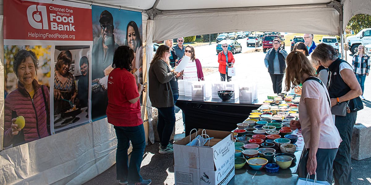 People Browse Colorful Bowls On Tables Under A Tent With A Food Bank Banner.