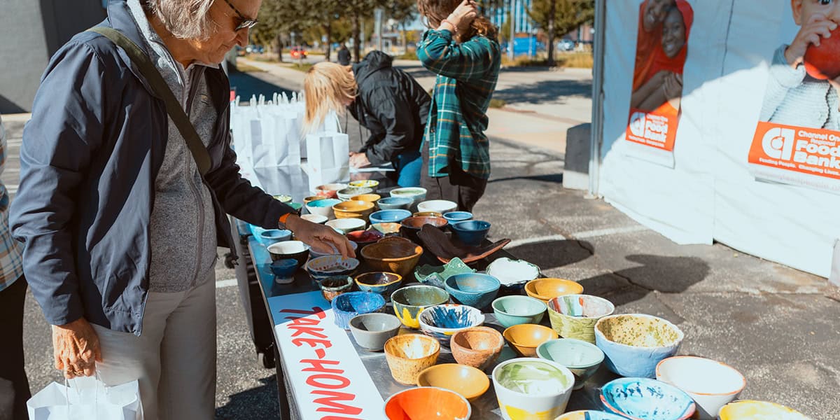 People Browse Colorful Ceramic Bowls On A Table At An Outdoor Event, With A Sign That Reads Take-Home.