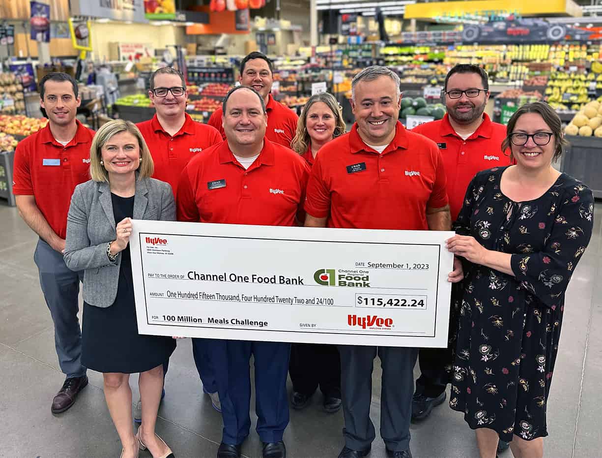 Hy-Vee 100 Million Meals Campaign Donation To Channel One Regional Food Bank_Photo Courtesy Of Hy-Vee A Group Of People Stands In A Grocery Store Holding An Oversized Check For $115,422.24 Made Out To Channel One Food Bank, Dated September 1, 2023.