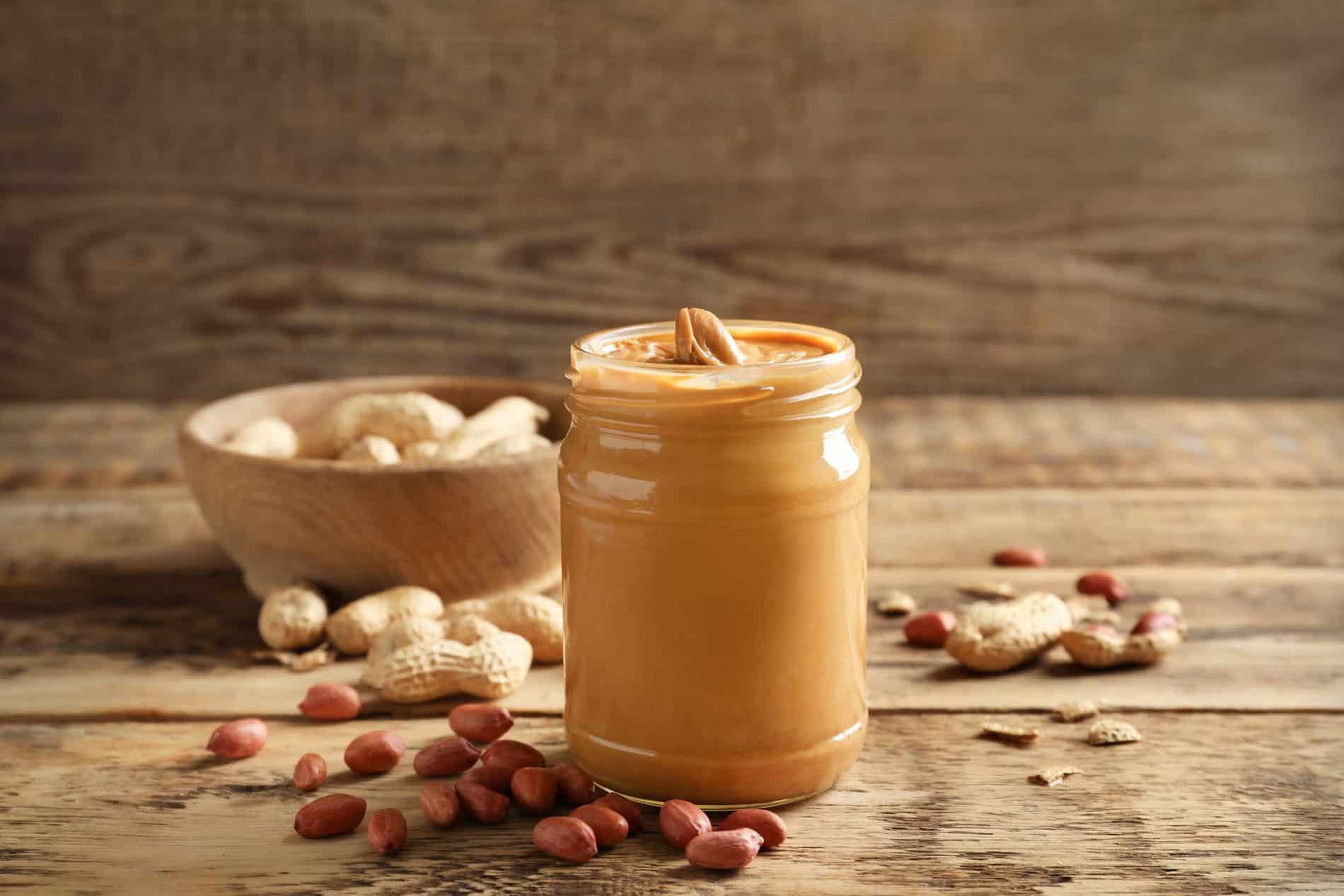 A Jar Of Peanut Butter Is On A Wooden Table Surrounded By Peanuts In Shells And A Wooden Bowl Filled With Peanuts.