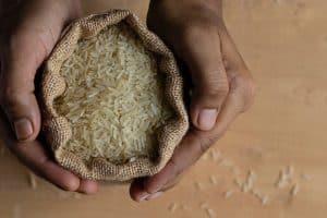Hands Holding A Burlap Sack Filled With Uncooked Rice On A Wooden Surface. Some Grains Are Scattered Outside The Sack.