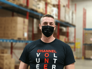 Channel-One-Individual-Volunteer A Man Wearing A Black Mask And A Volunteer T-Shirt Stands In A Warehouse With Shelves In The Background.